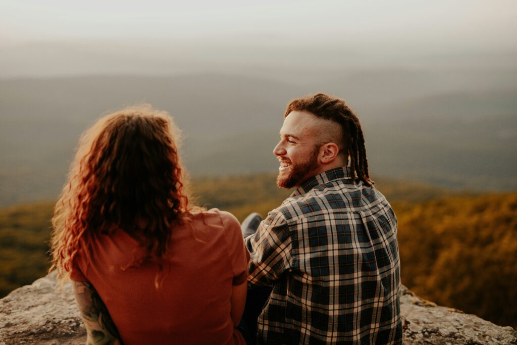 A couple sitting on a mountain ledge smiling at each other, symbolizing joy and connection fostered through therapy.