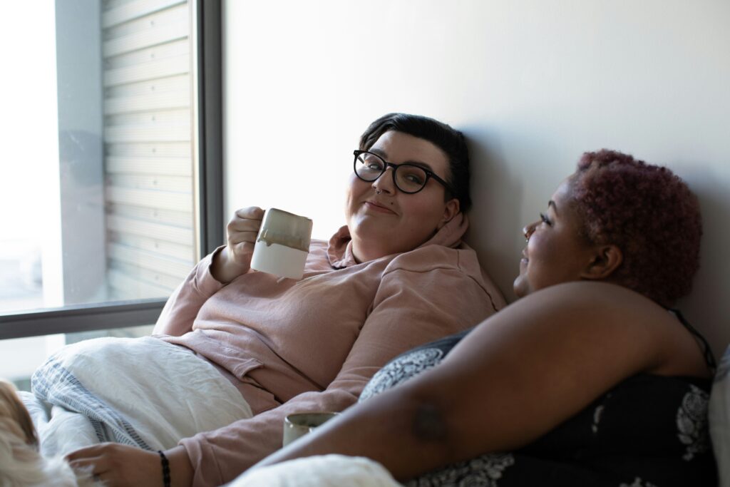 A couple cuddled up in bed by the window, drinking coffee and enjoying a peaceful, connected moment.