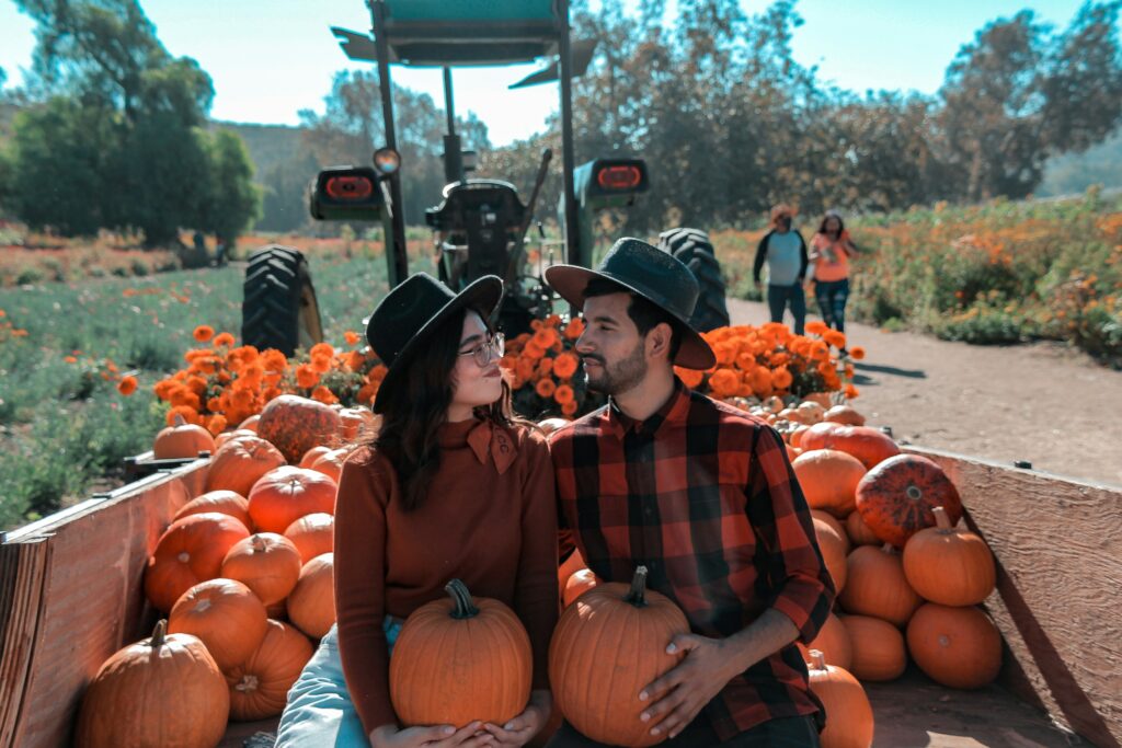 A couple sitting close in a pumpkin patch, holding pumpkins and smiling at each other, symbolizing warmth and connection during fall.