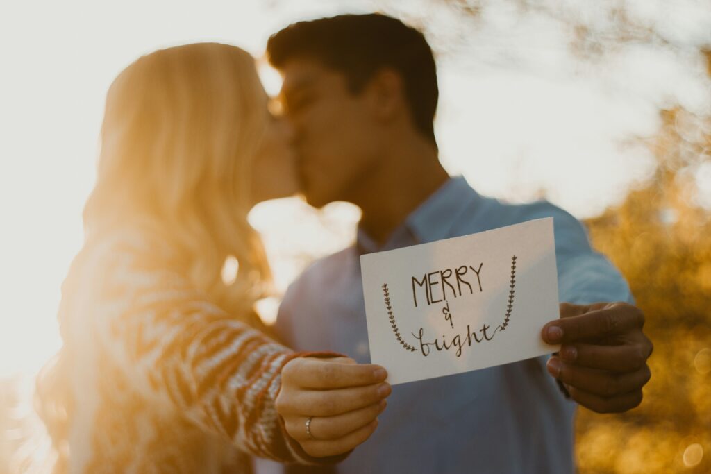 A couple kissing while holding a postcard that reads, "Merry and Bright"