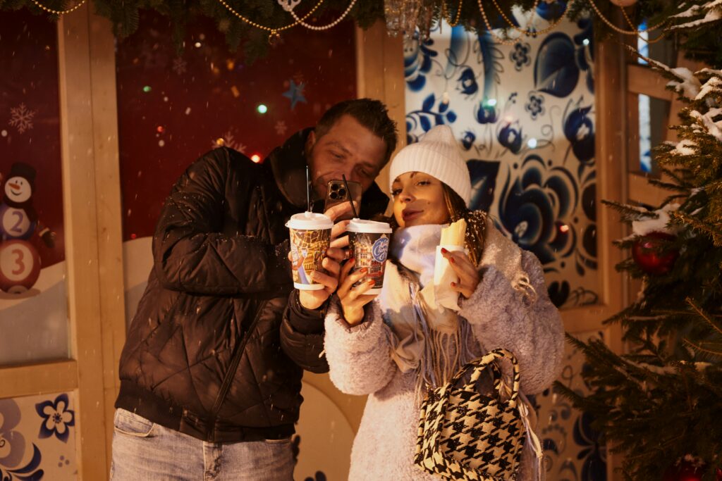 A couple bundled up at a festive winter market, enjoying hot drinks and a bite together, evoking cozy holiday traditions.