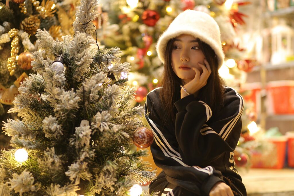 A young woman sitting by a Christmas tree, gazing thoughtfully, representing quiet holiday reflection.
